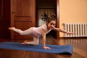 pequeno menina trabalhando Fora às lar, fazendo ioga exercício em uma ginástica esteira, ajoelhado oposto braço e perna extensão foto