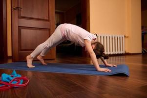 pequeno menina dentro roupa de esporte, alongamento dentro adho mukha svanasana exercício, para baixo voltado para cachorro pose, trabalhando Fora dentro de casa foto