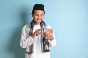 retrato do jovem ásia muçulmano homem segurando e tocante Móvel telefone com sorridente expressão em face. isolado imagem em azul fundo foto