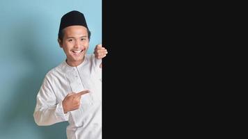 retrato do jovem ásia muçulmano homem mostrando produtos e apontando dedo às cópia de espaço em grande Preto papel poster publicidade seu texto. isolado imagem em azul fundo foto