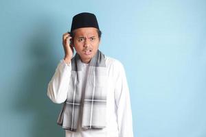 retrato do atraente ásia muçulmano homem dentro branco camisa pensando sobre questão com mão em têmpora. isolado imagem em azul fundo foto