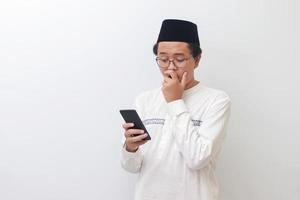 retrato do jovem ásia muçulmano homem segurando Móvel telefone e cobertura boca com chocado expressão em face. isolado imagem em branco fundo foto