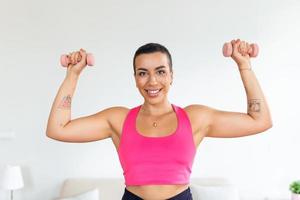 bíceps exercícios, braços dar certo. confiante Preto desportista fazendo Treinamento com dois azul halteres, bandeira, borrado fundo. retrato do sorridente Forte em forma senhora dentro branco roupa de esporte sutiã topo foto