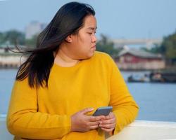 retrato gordo ásia mulher grandes Preto cabelo vestindo amarelo camisa estão usando Móvel telefone ou Smartphone trabalhos com sério e sério face e expressão dentro parque dentro tarde horas pôr do sol dia foto