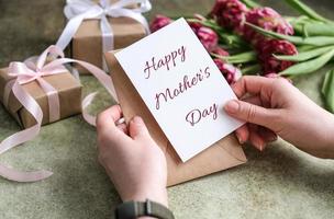 fechar-se do fêmea mãos segurando uma cartão com a texto feliz mãe dia em a fundo do uma ramalhete do tulipas e presentes. mãe dia conceito foto