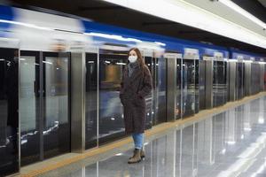 uma mulher com uma máscara facial de médico está esperando a chegada de um trem no metrô foto