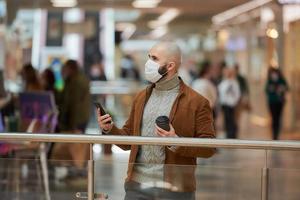 homem com máscara facial está usando um telefone e segurando um café no shopping foto