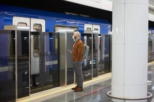 um homem com uma máscara facial segura um smartphone enquanto espera o trem do metrô foto