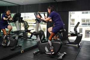 jovem fêmea com 1 protético perna com exercício com uma fiação bicicleta dentro a Academia para prática articulação usar com a de outros normal perna para estar flexível, conceito do vivo uma mulher vida com uma protético foto