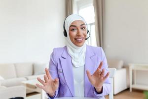 empresária dentro hijab tendo uma vídeo bate-papo em computador portátil . fêmea sentado às cafeteria e fazer vídeo ligar usando fones de ouvido e computador portátil computador. foto