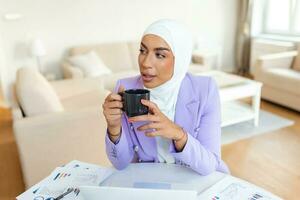 mulher muçulmana trabalhando com computador. mulher de negócios jovem árabe sentada em sua mesa em casa, trabalhando em um computador portátil e bebendo café ou chá. mulher muçulmana trabalhando em casa e usando o computador. foto