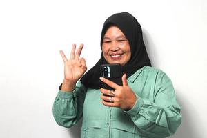 feliz meio envelhecido ásia mulheres vestindo hijab segurando Móvel telefone e mostrando Está bem gesto foto