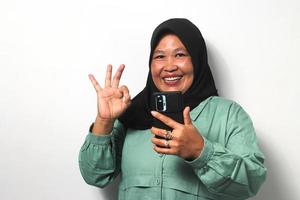 feliz meio envelhecido ásia mulheres vestindo hijab segurando Móvel telefone e mostrando Está bem gesto foto