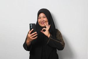 meio envelhecido ásia mulheres vestindo hijab segurando Móvel telefone e mostrando Está bem gesto foto