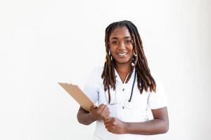 médico sorrisos dentro hospital corredor e segurando prancheta com paciente arquivo. africano americano fêmea pediatra enfermeira dentro escritório. retrato do fêmea enfermeira em pé dentro hospital corredor foto