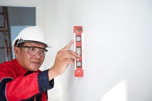 uma construção trabalhador ou arquiteto vestindo uma Difícil chapéu é verificação a parede com uma nível medidor. foto