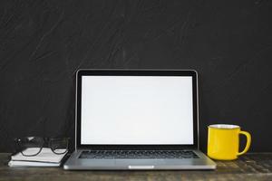 laptop e óculos com caneca de café amarela e diário na mesa