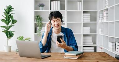 sorridente masculino relaxante às lar, ela é jogando música usando Smartphone tábua, computador portátil, e vestindo branco fones de ouvido. em mesa foto