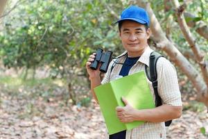 ásia homem explorador desgasta azul boné, detém binocular dentro floresta para pesquisa botânico plantas e criaturas animais selvagens. conceito, natureza exploração. ecologia e ambiente. foto