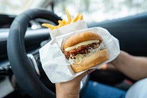 ásia senhora segurando Hamburger e francês fritas para comer dentro carro, perigoso e risco a acidente. foto