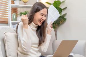 feliz excitado, ásia jovem mulher segurando papelada ou documento recebendo, recebido trabalho promoção, aprovar imposto reembolso ou empréstimo hipoteca, olhando Boa notícia às computador portátil computador, sentado em sofá às lar. foto