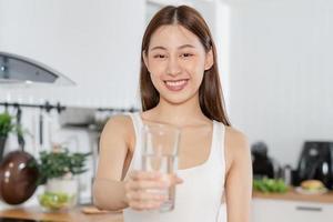 feliz lindo, ásia jovem mulher, menina bebendo, trago fresco vidro do água para hidrato, segurando transparente vidro dentro dela mão, com sede às lar. saúde Cuidado, saudável estilo de vida conceito. foto