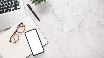 branco escritório escrivaninha local de trabalho com em branco tela Smartphone, caderno, óculos, computador portátil e caneta, topo Visão plano deitar com cópia de espaço. foto