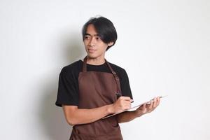 retrato do atraente ásia barista homem dentro Castanho avental levando ordem, escrevendo em cardápio livro lista. isolado imagem em branco fundo foto