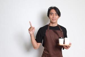 retrato do atraente ásia barista homem dentro Castanho avental segurando levar longe papel café copo com de madeira bandeja para servir enquanto apontando para a lado. isolado imagem em branco fundo foto