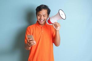 retrato do atraente ásia homem dentro laranja camisa Falando mais alto usando megafone, promovendo produtos enquanto segurando Móvel telefone. publicidade conceito. isolado imagem em azul fundo foto