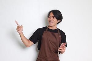 retrato do atraente ásia barista homem dentro Castanho avental segurando e apontando Móvel telefone. isolado imagem em branco fundo foto
