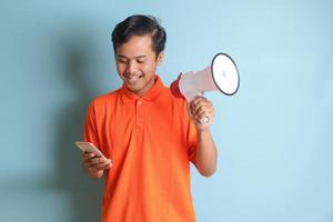 retrato do atraente ásia homem dentro laranja camisa Falando mais alto usando megafone, promovendo produtos enquanto segurando Móvel telefone. publicidade conceito. isolado imagem em azul fundo foto