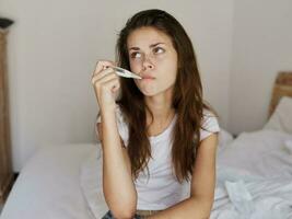 mulher esperando com uma termômetro dentro dela boca sentado em a cama foto