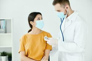 médico dentro médico mascarar e casaco examinando mulher dentro amarelo camiseta foto