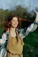 retrato do uma lindo jovem mulher com vermelho cabelo dentro natureza dentro uma parque dentro a verão, olhando às a pôr do sol contra uma fundo do verde Relva e árvores foto