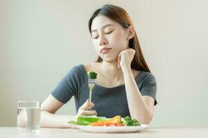 dieta dentro entediado, infeliz lindo ásia jovem mulher, menina em fazer dieta, segurando garfo às brócolis dentro salada placa, não gostar ou cansado com comer fresco legumes. nutrição do limpar, saudável Comida Boa gosto. foto