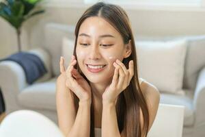 fresco saudável pele, lindo sorrir do ásia jovem mulher, menina olhando às espelho, aplicando hidratante em dela face, colocando creme tratamento antes Maquiagem Cosmético rotina às lar. facial beleza. foto