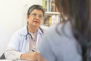 maduro, Senior ásia médico, psiquiatra, geral mulher médico consultando preocupado com jovem paciente, usando estetoscópio para ouvir, compromisso às clínica, hospital. saúde Cuidado, Verifica acima médico. foto