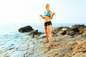 atleta mulher fazendo esporte exercício com a corda em a pedras de a mar foto
