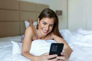 sorridente mulher olhando às Móvel telefone enquanto deitado em branco cama. feliz morena jovem mulher usando celular às lar. lindo menina surfar a internet em Smartphone foto