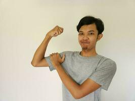 animado ásia homem vestindo uma cinzento camisa mostrando Forte gesto de elevação dele braços e músculos sorridente orgulhosamente foto