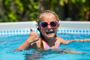 fofa pequeno menina sorridente dentro oculos de sol dentro a piscina em uma ensolarado dia. a criança mostrando polegares acima gostar gesto olhando Câmera . verão período de férias foto