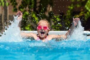 fofa pequeno menina sorridente dentro óculos dentro a piscina em uma ensolarado dia. a criança espirrando água dentro a piscina . verão período de férias foto