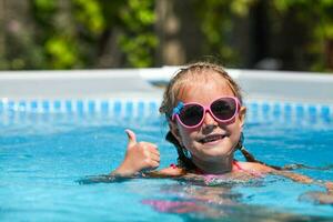 fofa pequeno menina sorridente dentro oculos de sol dentro a piscina em uma ensolarado dia. a criança mostrando polegares acima gostar gesto olhando Câmera . verão período de férias foto