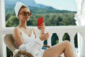 atraente jovem mulher desfrutando a manhã em a sacada olhando às a Móvel telefone tela montanha Visão foto