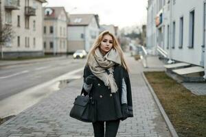 lindo elegante jovem mulher dentro a outono casaco com uma cachecol com uma Bolsa caminhando em uma cidade foto