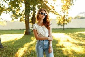 lindo jovem menina dentro uma branco camiseta e vintage jeans com oculos de sol dentro a parque em uma ensolarado dia. foto