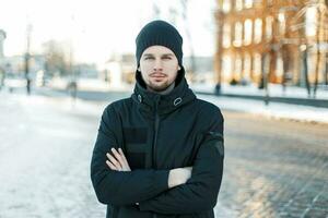 bonito jovem homem dentro uma tricotado boné e uma Preto Jaqueta em uma inverno dia às pôr do sol foto