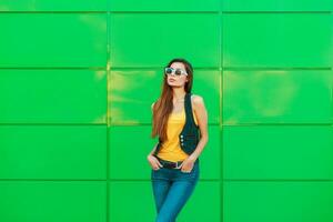 lindo mulher dentro uma amarelo camiseta e jeans Jaqueta em pé perto uma verde metal muro. foto