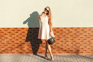 lindo mulher dentro uma Rosa vestir com Bolsa em uma ensolarado dia em pé perto a parede foto